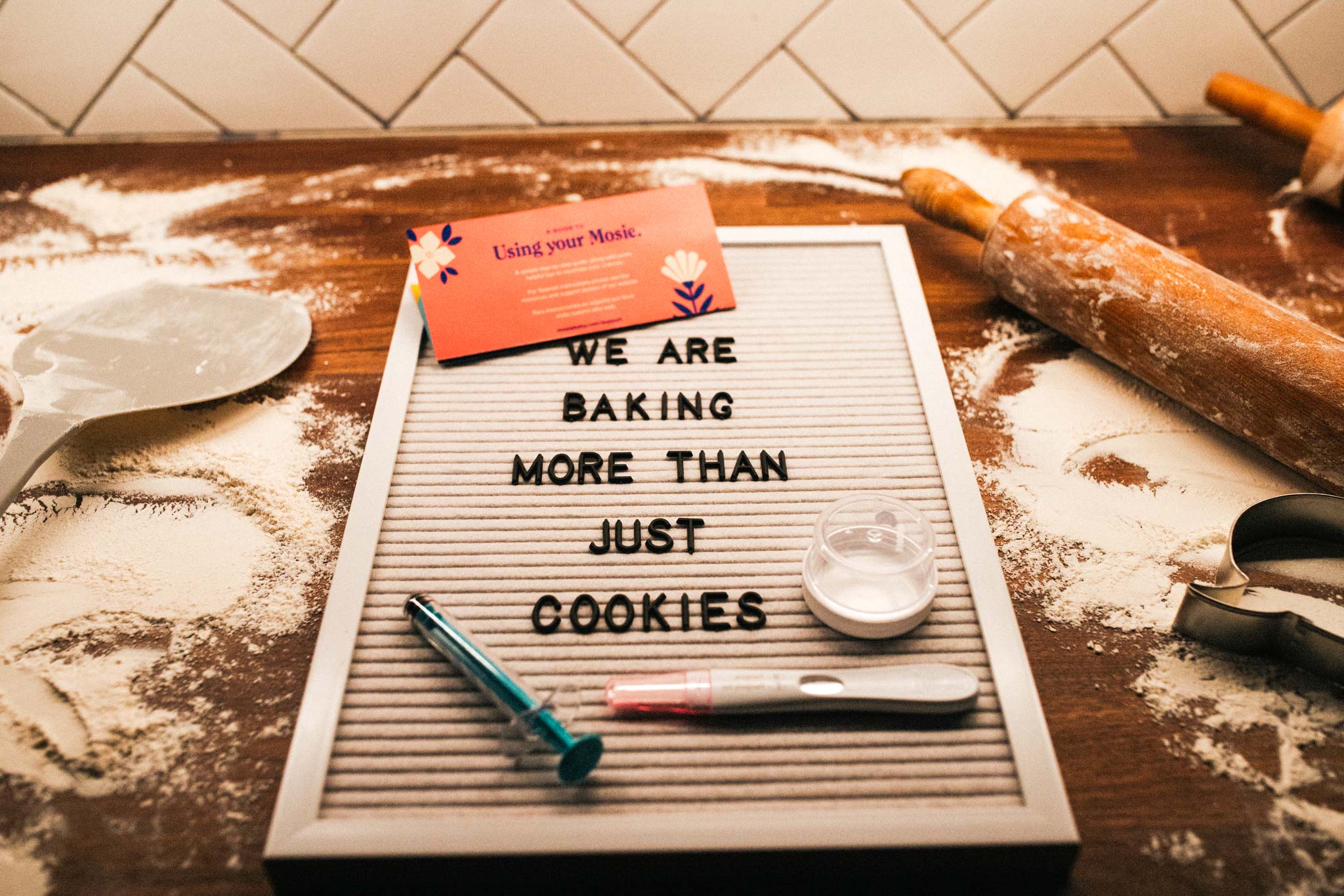Mosie Baby announcement on letterboard that reads "We are baking more than just cookies" on flour covered counter with baking tools, positive Mosie Baby pregnancy test and Mosie Baby Kit.