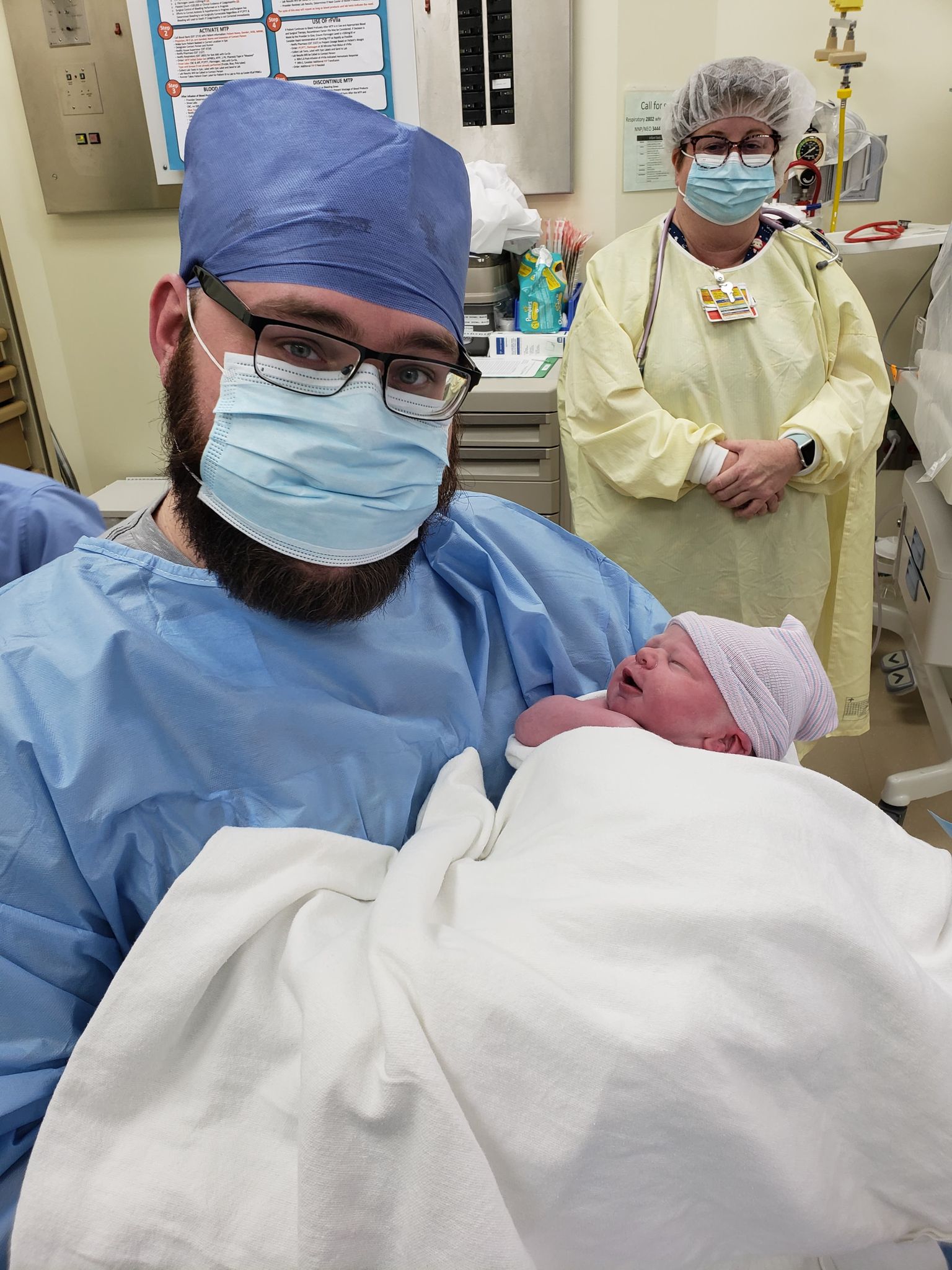 Man in blue surgical mask and scrubs holds newborn with doctor standing in background