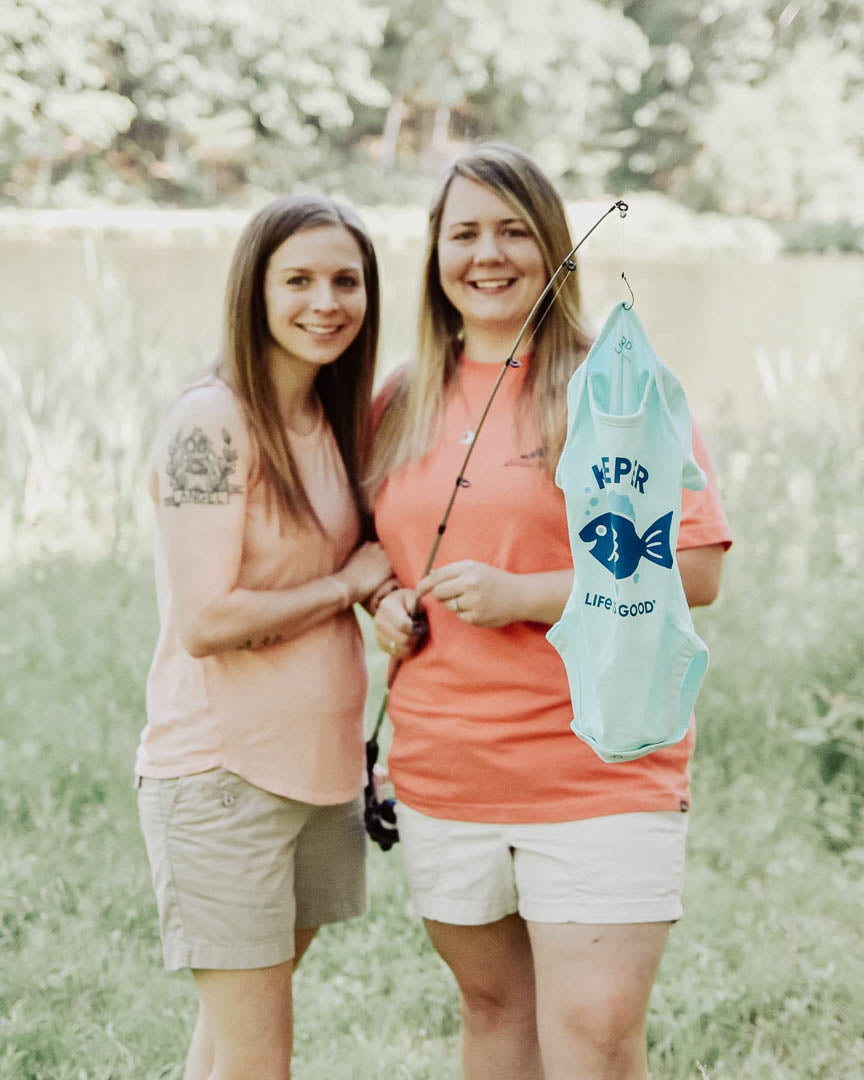 Mamas-to-be share pregnancy announcement outside with fishing rod with a onesie at the end of it.