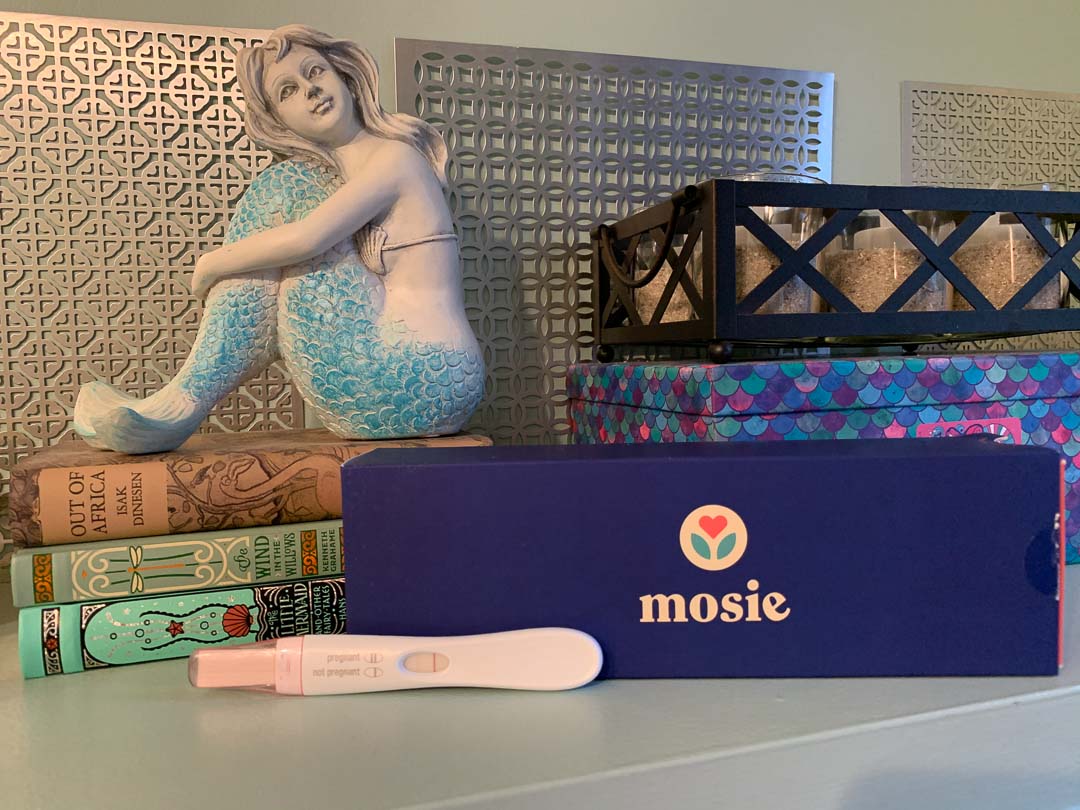 Positive pregnancy test and The Mosie Kit box on side table in someone's home.