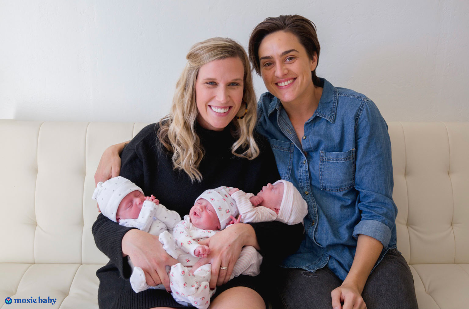An LGBTQ couple sit on the couch holding triplets