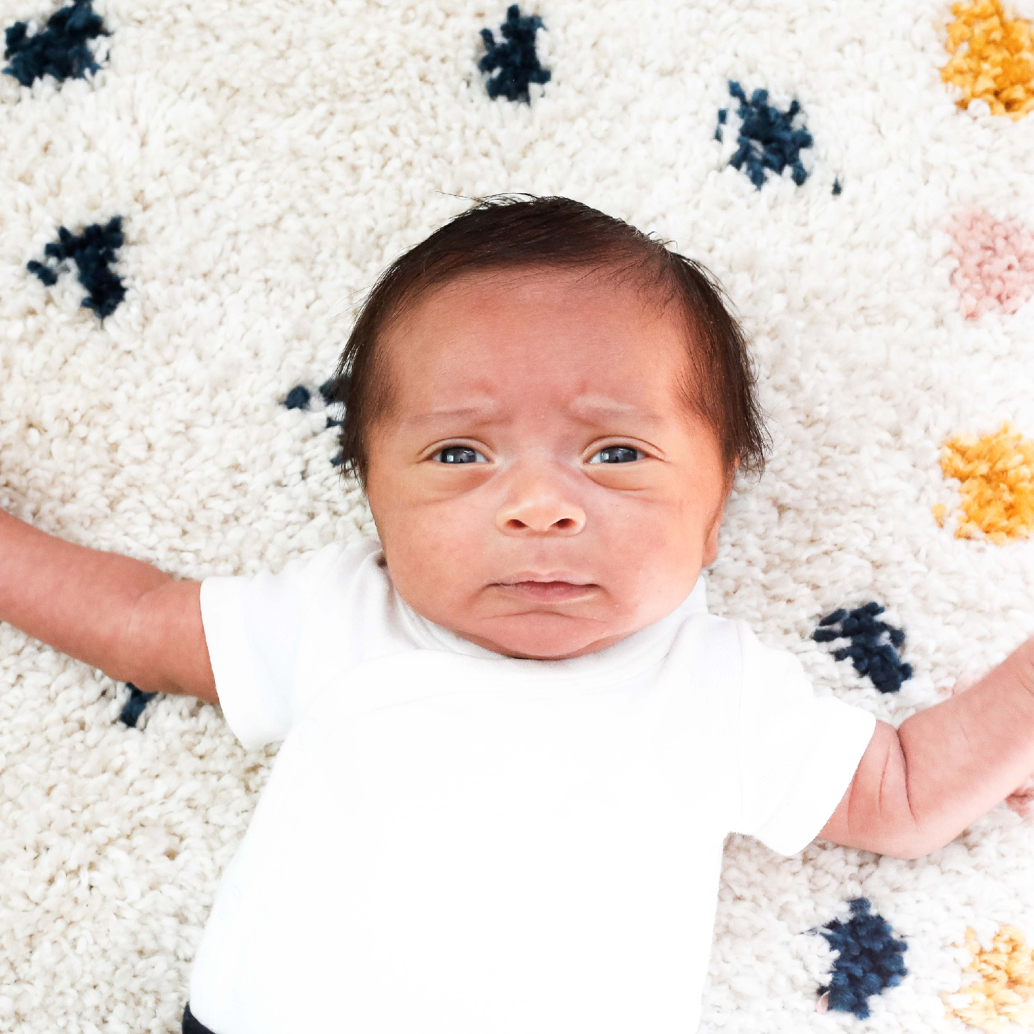 Newborn baby boy with dark hair and white onesie lays on white rug with blue and yellow polka dots.