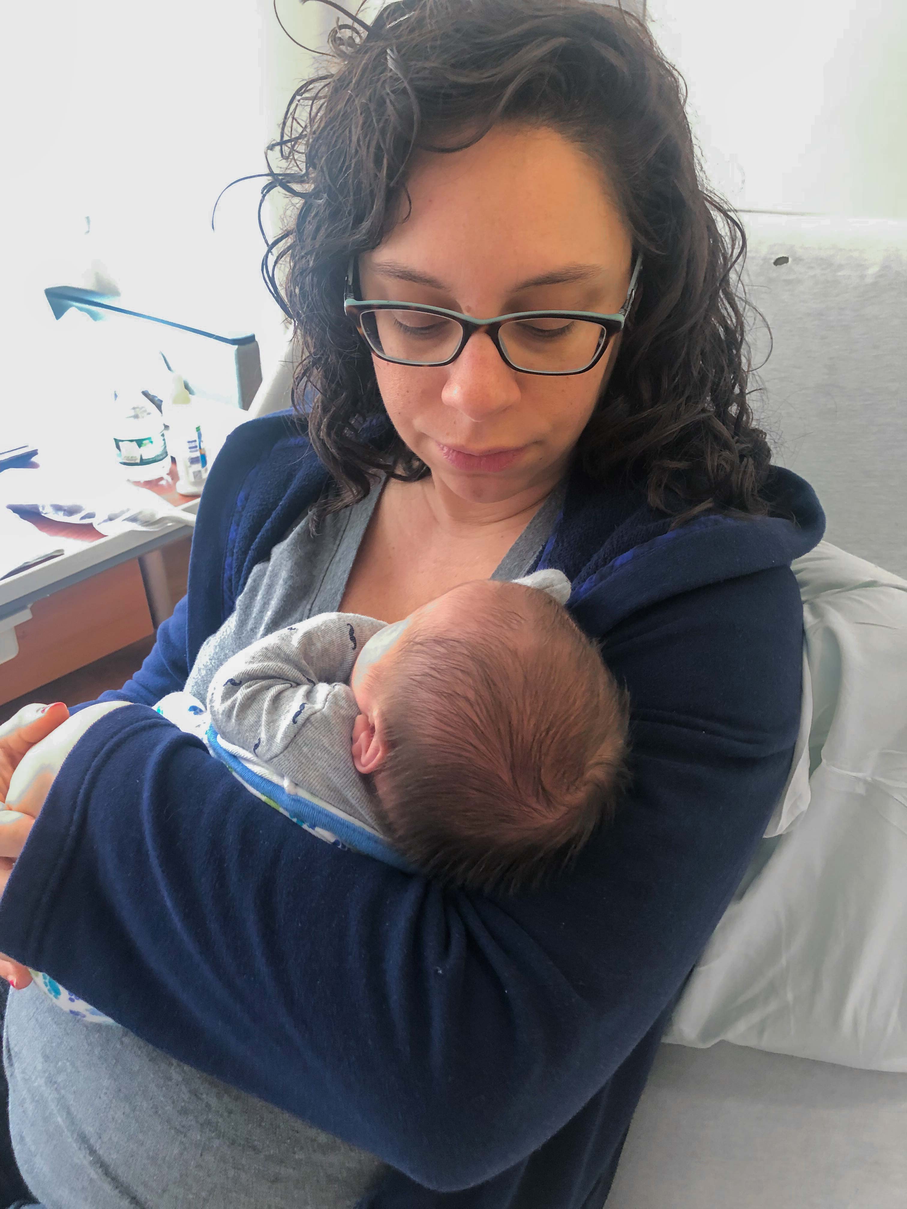 New mama holds her Mosie Baby in her arms with black glasses and curly dark hair.