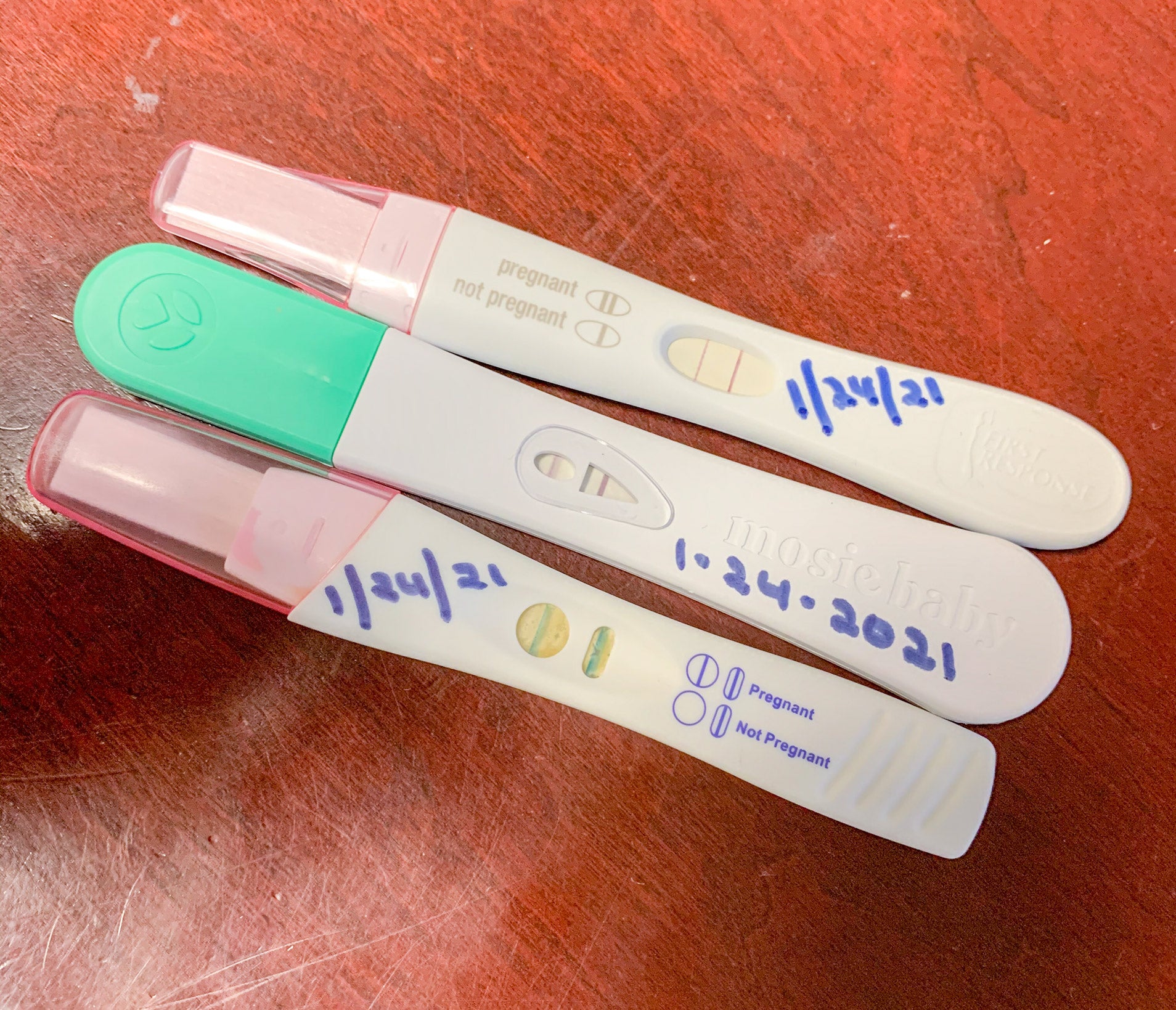 Three positive pregnancy tests, two pink ones and a Mosie pregnancy test with dates written in marker on top of each.
