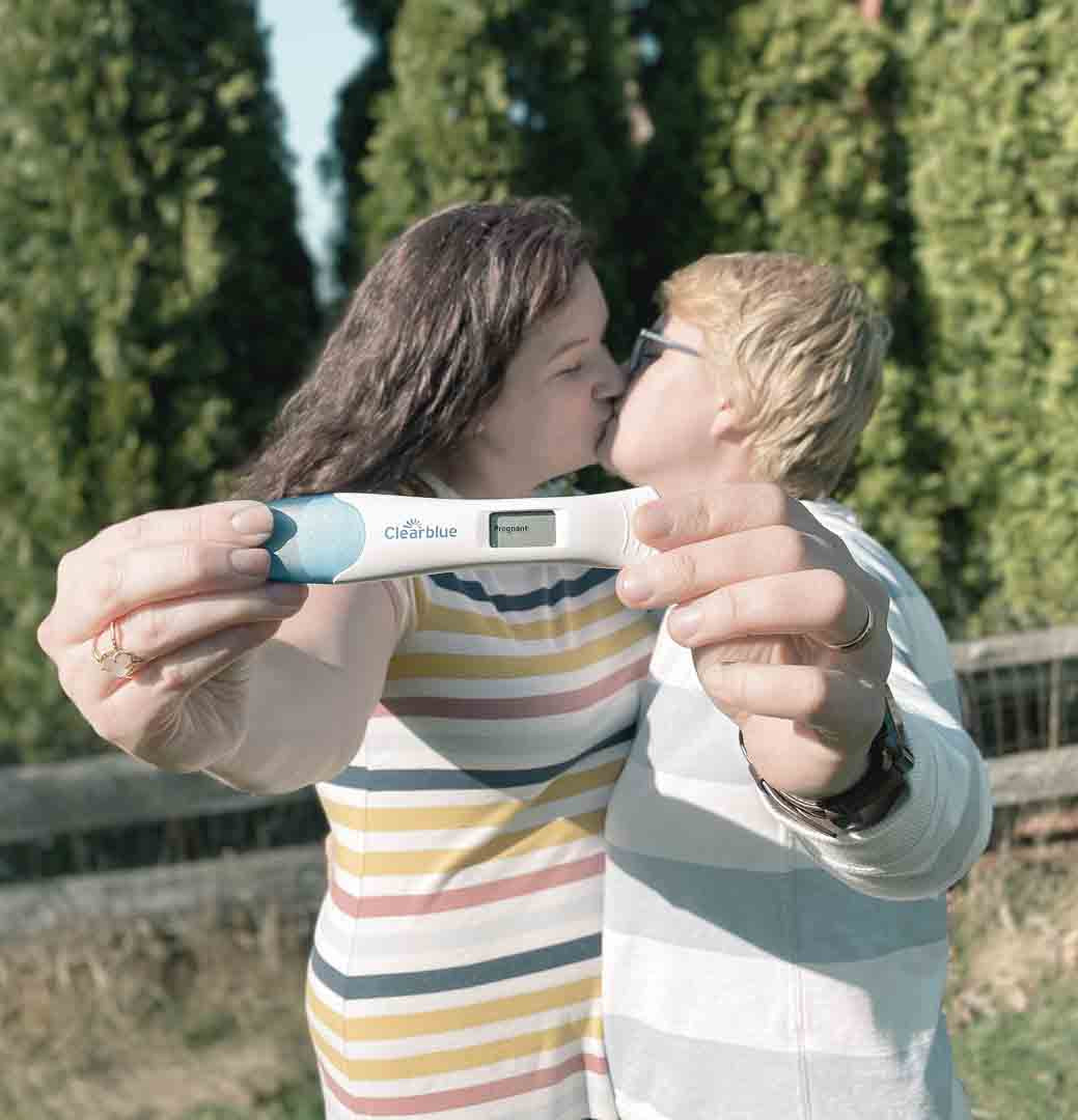Married women kiss outside while holding positive pregnancy test together towards camera.