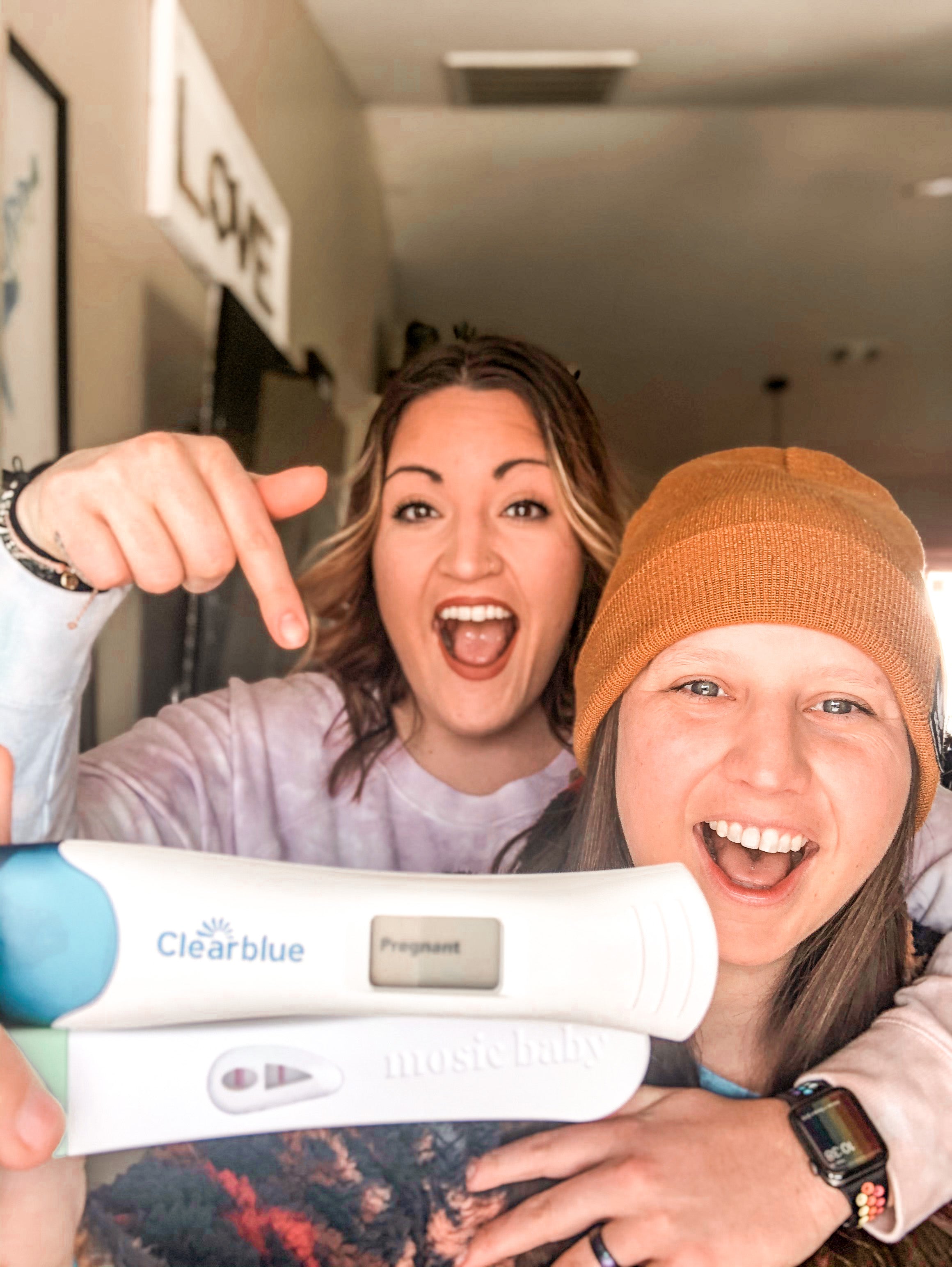 Mama's-to-be hold up two positive pregnancy tests, with wife on the left pointing and smiling to the tests as the wife on the right holds them.