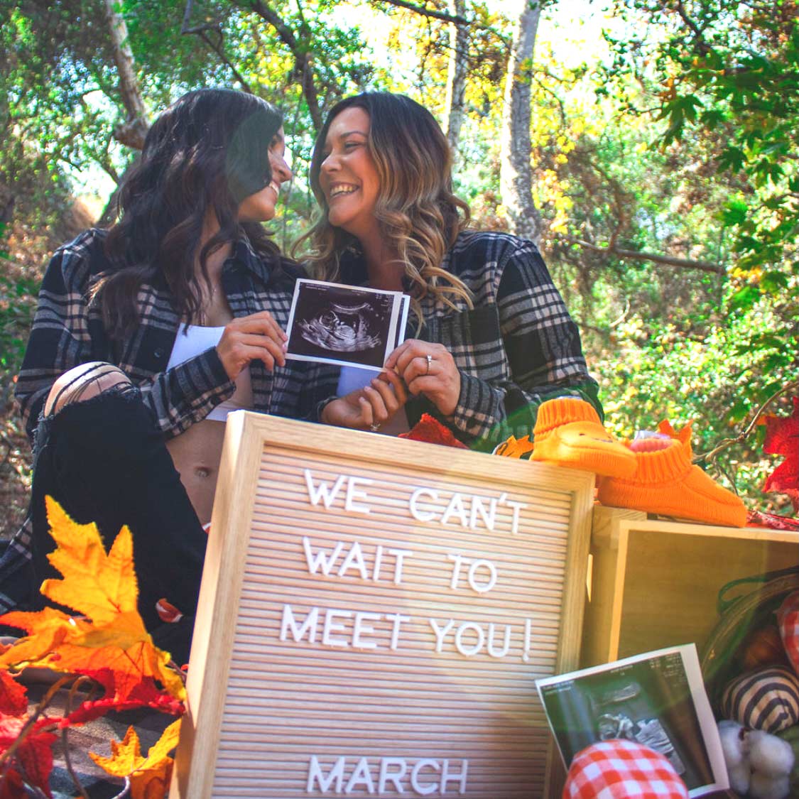 Lesbian couple holding an ultrasound of their mosie baby in the fall with a sign that says "we can't wait to meet you, March"