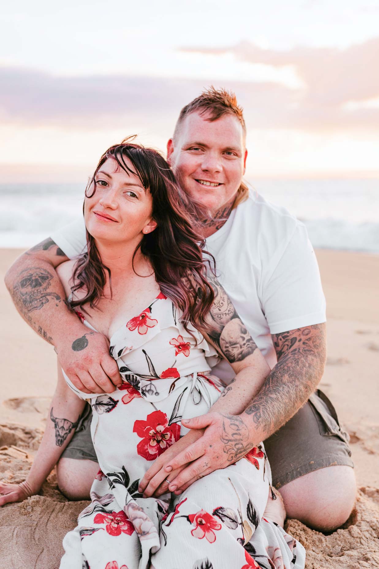 Man in white shirt and gray shorts holds pregnant wife in white dress with red flowers from behind lovingly on an Australian beach