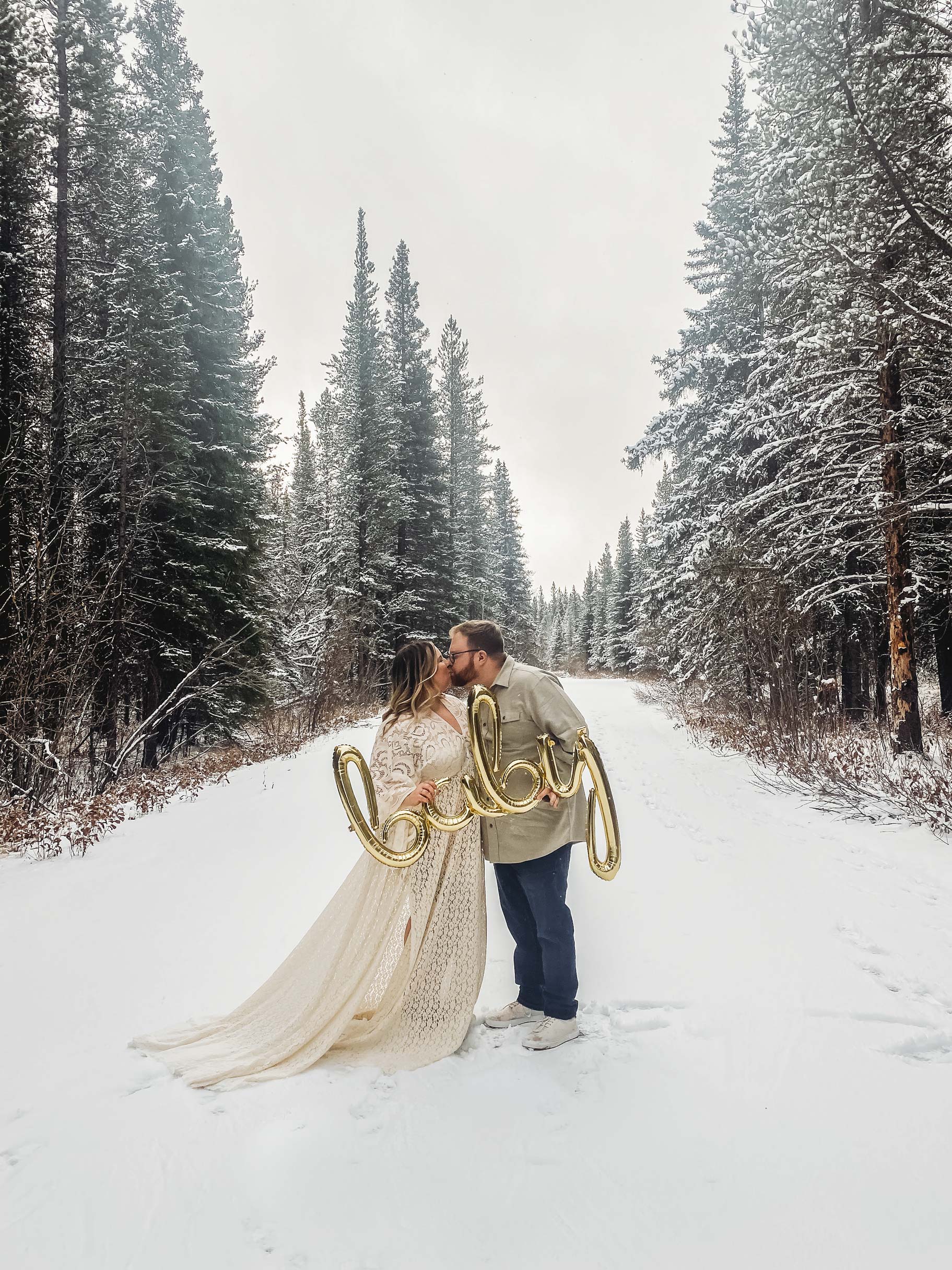 Canadian Husband and wife kiss while holding giant balloons that spell out the word 'baby' standing in a winter wonderland of snow and fir trees