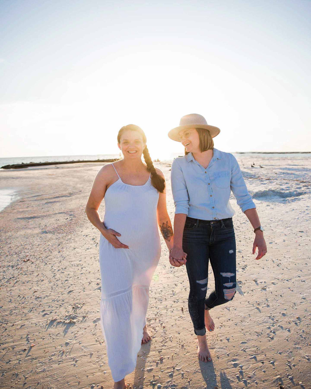 Pregnant woman in white dress holds her wife's hand on the beach.