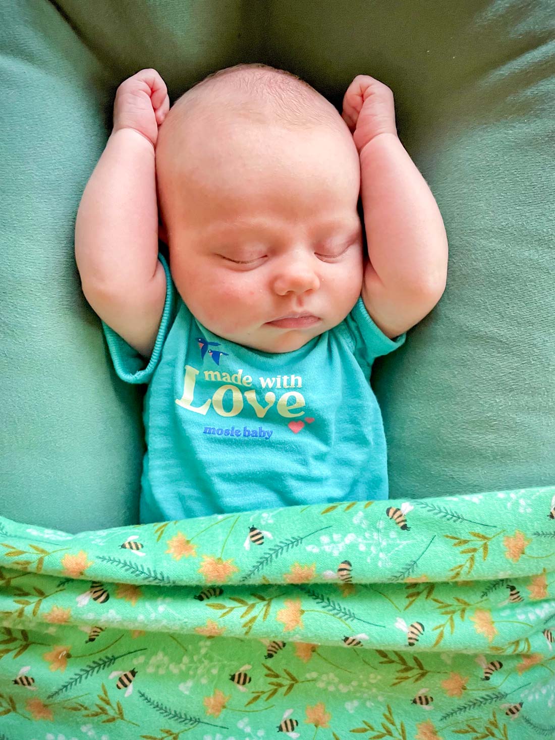 Newborn baby sleeps cozily in Mosie Baby onesie and big pillow with green blanket