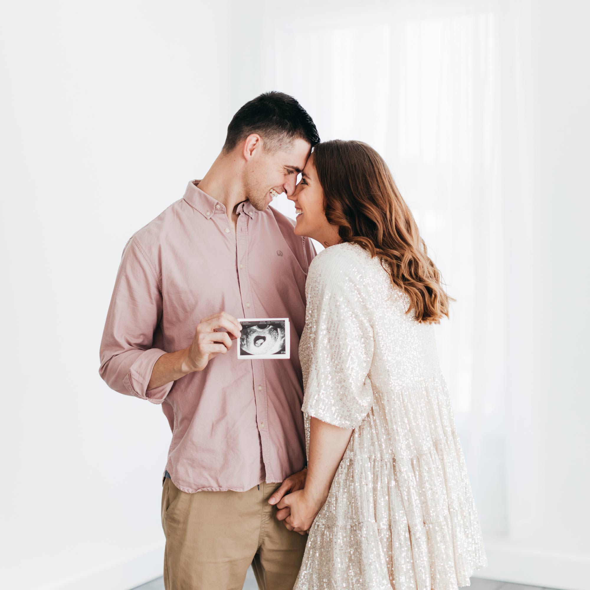 couple standing in front of rainbow heart banner holding a sonogram