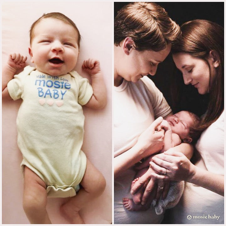 A baby in a Mosie baby onesie being help by two moms 